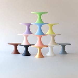 Puede incluir: Una colección de pequeñas mesas de pedestal en varios colores pastel y metálicos. Las mesas están dispuestas en forma de pirámide, mostrando una gama de tonos que incluyen verde, azul, rosa y dorado. Cada mesa tiene una parte superior redonda y una base acampanada.