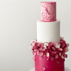 Puede incluir: Un pastel de boda de tres pisos con un piso superior de mármol rosa, un piso blanco con líneas verticales y un piso inferior con glaseado rosa oscuro y pétalos de flores rosas y blancas.