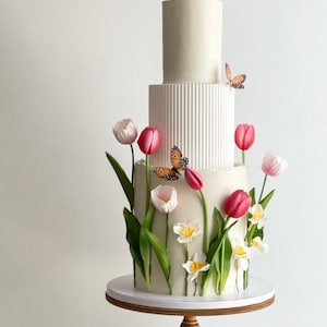May include: A white wedding cake decorated with fondant tulips and butterflies. The cake has three tiers, with the bottom tier being the largest and the top tier being the smallest. The cake is decorated with white fondant and has a smooth finish. The tulips are pink and white, and the butterflies are orange and brown.