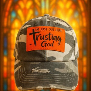 May include: Camouflage baseball cap with an orange patch that reads "I'M JUST OUT HERE Trusting God" in black script. The cap has a distressed brim and a classic six-panel design. The background is a blurred stained-glass window.