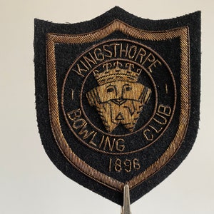 May include: A black felt patch with gold embroidered lettering and a crest. The patch reads "Kingsthorpe Bowling Club 1896".