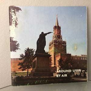 Op de afbeelding: Een vintage reisposter met een rood bakstenen gebouw met een klokkentoren en een standbeeld van een man die naar het gebouw wijst. De tekst op de poster luidt "Around USSR by Air".