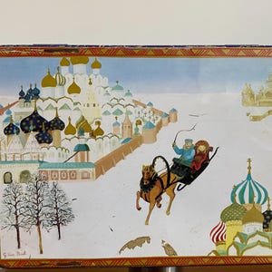 Può includere: Una scatola di latta vintage con un'illustrazione colorata di una scena di città russa innevata. La scatola presenta una slitta trainata da un cavallo con due persone a bordo, e un gruppo di edifici con cupole a forma di cipolla. Il testo "G.Ivanov. Brest" è stampato nell'angolo in basso a sinistra.
