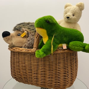 Op de afbeelding: Een rieten mand gevuld met knuffels, waaronder een bruine egel, een groene kikker en een witte teddybeer.
