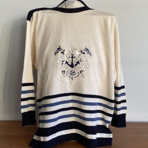 May include: Cream-coloured sweater with navy blue and light blue stripes at the bottom and on the sleeves. The back features an embroidered nautical crest with an anchor, flags, and ornate detailing. The sweater has a classic, preppy style.