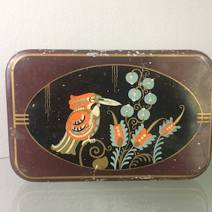 Puede incluir: Una lata de metal vintage con fondo marrón oscuro y un diseño ovalado negro. El óvalo presenta un pájaro estilizado con plumas naranja, amarillo y negro, rodeado de flores azules y verdes.