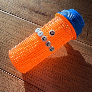 May include: An orange pill bottle with a blue cap. The word "PILLS" is spelled out in white letters on the side, with a smiley face. The bottle is on a wooden surface.