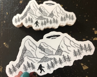 May include: Black and white sticker featuring a mountain range with a Bigfoot and a UFO. The sticker is on a white background.