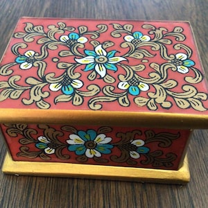 May include: A small wooden box with a red base and gold trim. The top and bottom of the box are decorated with a floral pattern in gold, white, and blue.