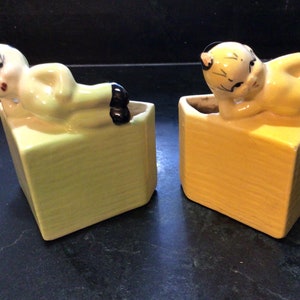 May include: Two ceramic planter figurines, one yellow and one green, each depicting a stylized person lying down. The planters are shaped like a cube with a cut-out section for planting.
