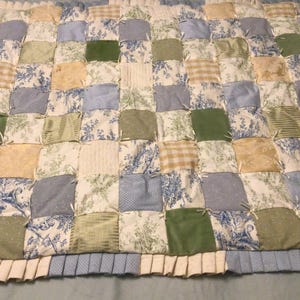May include: A handmade patchwork quilt featuring a variety of fabrics in shades of blue, green, cream, and yellow. The quilt has a ruffled edge and is made of square patches with floral, striped, and solid patterns.