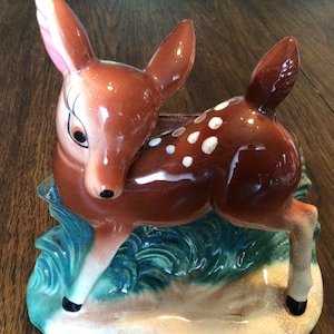 May include: A ceramic deer figurine planter in shades of brown, white, and blue. The deer has white spots and is posed on a base with green and blue foliage. The planter is a vintage decorative item.