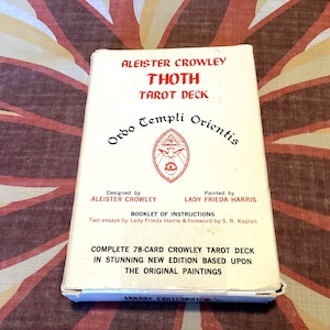 May include: A white rectangular box for the Aleister Crowley Thoth Tarot Deck. The front features red and black text, including the title, Ordo Templi Orientis, and credits to Aleister Crowley and Lady Frieda Harris, plus a central red emblem.