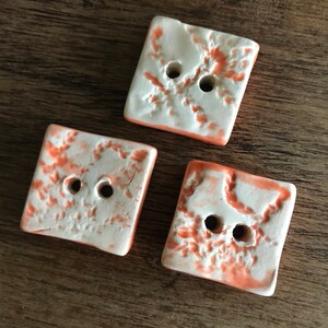 May include: Three square ceramic buttons with a white background and a light orange design. The buttons have two holes each.