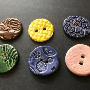Könnte beinhalten: Sechs Keramikknöpfe mit verschiedenen Designs und Farben. Die Knöpfe sind rund und haben jeweils zwei Löcher. Die Designs umfassen ein gewebtes Muster, ein Blumenmuster, ein Blattmuster und ein geometrisches Muster. Die Farben umfassen Gelb, Blau, Grün, Braun und Rosa.