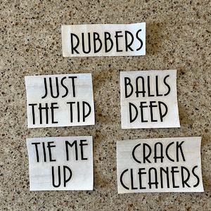 May include: Six white paper squares with black text. The text reads "RUBBERS", "JUST THE TIP", "BALLS DEEP", "TIE ME UP", "CRACK CLEANERS".