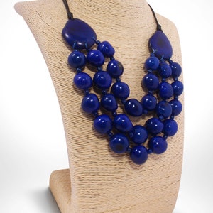May include: A three-strand necklace made of large, dark blue, round beads. The necklace is displayed on a beige, woven mannequin bust.