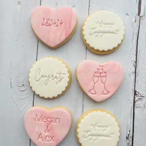 May include: Six decorated sugar cookies, two are heart-shaped with pink marbling, two are round with white icing and scalloped edges, and two are round with white icing. The cookies are decorated with pink icing and have various messages including "Congrats", "Happy Engagement", "Megan & Alex", and "280424".