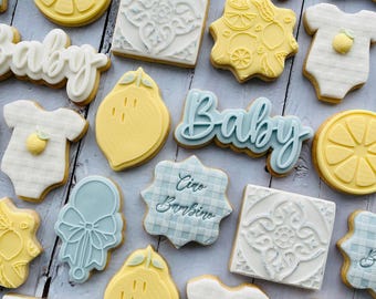 May include: A collection of decorated sugar cookies featuring yellow lemon slices, white onesies with small yellow lemons, light blue rattles, square tiles with white embossed patterns, and cookies with the text Baby or Ciao Bambino in script.