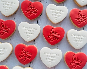 May include: Heart-shaped cookies with red and white icing. Red cookies say "Love you" with an arrow, while white cookies say "Consider this a hug in a biscuit". A sweet treat for Valentine's Day.