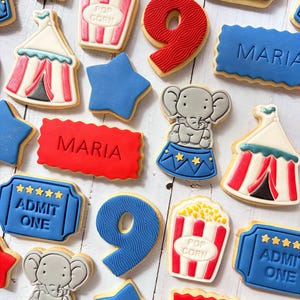 May include: A collection of circus-themed sugar cookies featuring red and blue colours. Shapes include popcorn buckets, circus tents, elephants on pedestals, stars, the number 9, and rectangular plaques with the text ADMIT ONE or MARIA.