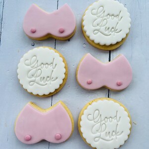 May include: Six decorated sugar cookies. Three cookies are pink and shaped like a bra. Three cookies are white with the text "Good Luck" in cursive.