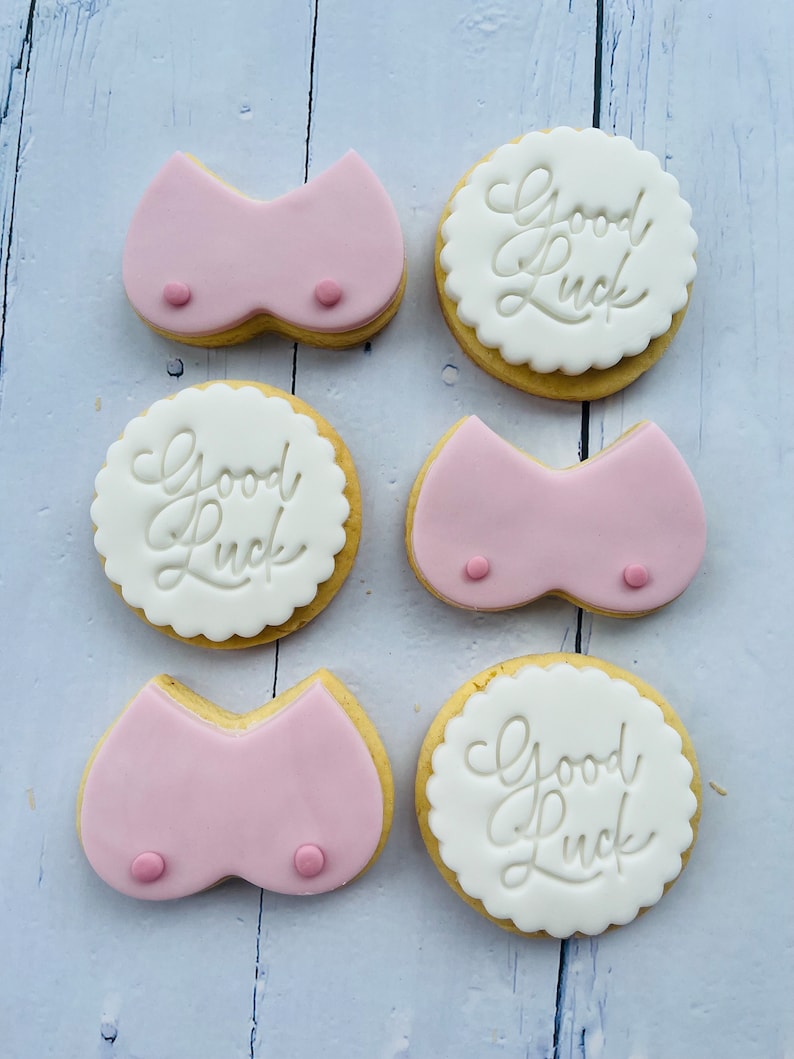 May include: Six decorated sugar cookies. Three cookies are pink and shaped like a bra. Three cookies are white with the text "Good Luck" in cursive.