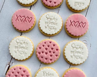 May include: Overhead shot of a dozen round cookies with white and pink frosting. Some cookies have the text "Fries before guys" or "Sisters before Mistas". Others have heart patterns or "XOXO" text.