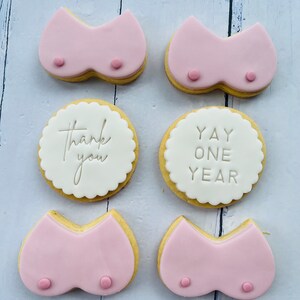 May include: Six decorated sugar cookies. Two cookies are pink and shaped like a bra. Two cookies are white with scalloped edges and say "Thank you" and "Yay One Year". Two cookies are pink and shaped like a bra.