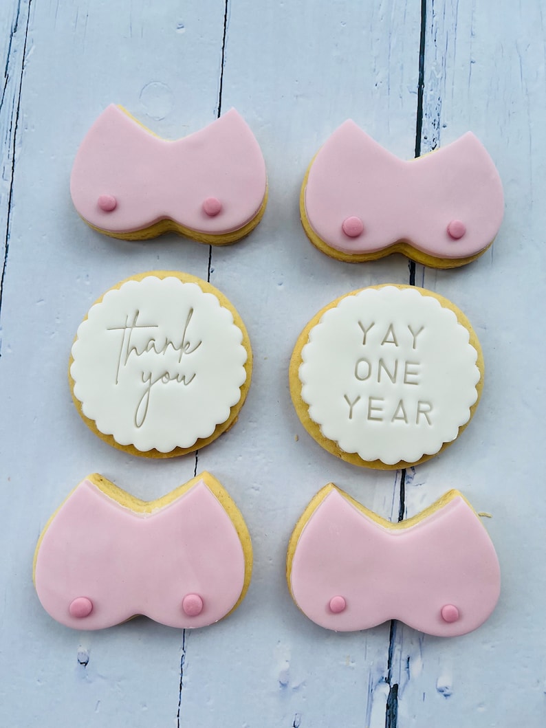 May include: Six decorated sugar cookies. Two cookies are pink and shaped like a bra. Two cookies are white with scalloped edges and say "Thank you" and "Yay One Year". Two cookies are pink and shaped like a bra.
