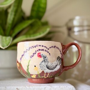 May include: Cream ceramic mug with a brown handle and base. The mug has a hand-painted design of a hen, chick, and lavender sprigs. The mug is set against a blurred background with a plant and a glass jar.