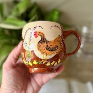 May include: A ceramic mug with a hand-painted design featuring a brown hen with a red comb and yellow chicks. The mug has a brown handle and base, with a cream-colored background and floral accents. The mug is held in a hand.