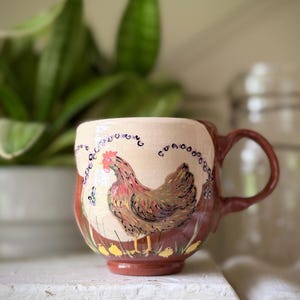 Peut inclure: Mug en céramique avec un motif de poule peint à la main. La tasse a une anse et une base marron, avec une partie supérieure crème. La poule est représentée en nuances de marron, rouge et jaune, avec de petits poussins jaunes à la base.