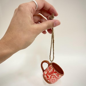 May include: A small, ceramic mug with a brown handle and rim, suspended by a jute string. The mug features a white base with a red floral pattern. The mug is held against a white background.