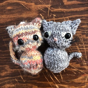 Pode incluir: Dois brinquedos de gato de malha, um com um padrão listrado cinza e branco e o outro com um padrão salpicado cinza e branco. Ambos têm olhos pretos e narizes cor de rosa.