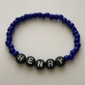 Pu&ograve; includere: Un braccialetto di perline blu con perline a forma di lettera nere che compongono il nome "HENRY".