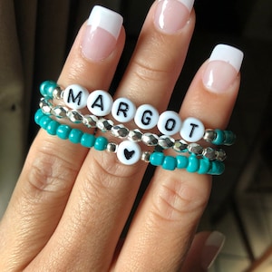 May include: A set of three turquoise beaded bracelets with a silver beaded bracelet and a white heart charm. The silver bracelet has white letter beads spelling out the name "MARGOT".