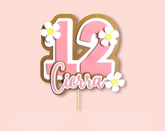 May include: A pink and white layered cake topper with the number 12 and the name "Cierra" in cursive. The topper is decorated with white daisies with yellow centers and has a wooden stick for insertion.