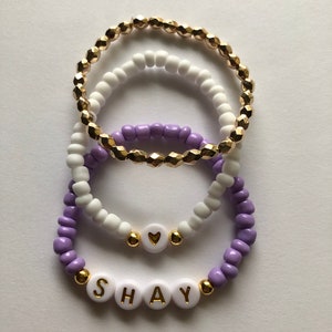 May include: Three beaded bracelets, one purple, one white, and one gold. The purple and white bracelets have a heart charm and the purple bracelet has the name "SHAY" spelled out in white beads.