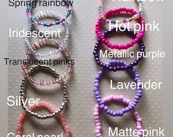 May include: A set of ten colorful beaded bracelets, including rainbow, hot pink, iridescent, metallic purple, translucent pinks, lavender, silver, and coral pearl. The bracelets are arranged vertically in a row, with the names of each color written below them.