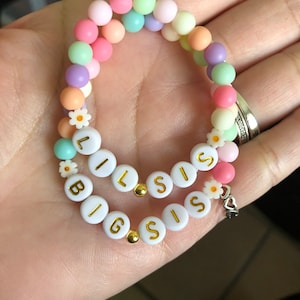 May include: A pastel-colored beaded bracelet with white beads spelling out "BIG SIS" and "Lil SIS". The bracelet has small white flowers and gold accents.