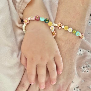 May include: Two people wearing personalized beaded bracelets with flower accents. The bracelets spell out the names "Casey" and "Mama".
