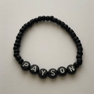 Pu&ograve; includere: Braccialetto di perline nere con il nome "PAYSON" scritto in lettere bianche.