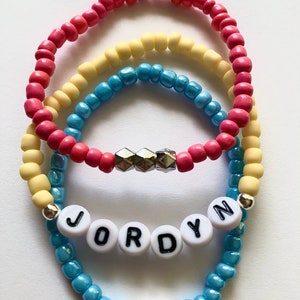 May include: Three layered bracelets with colorful beads. The top layer is pink, the middle layer is yellow, and the bottom layer is blue. The blue layer has a silver heart charm and white beads spelling out the name "Jordyn".