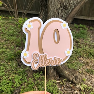 May include: A pink and brown cake topper with the number 10 and the name "Ellerie" in a cursive font. The number is decorated with white daisies.