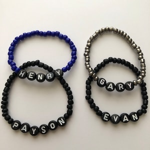 Pu&ograve; includere: Tre braccialetti di perline nere con perline lettere bianche che compongono i nomi "Payson", "Henry" e "Evan". Un braccialetto di perline argentate con perline lettere nere che compongono il nome "Gary" &egrave; sovrapposto ai braccialetti neri.