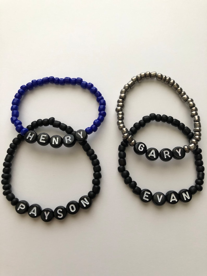 Pu&ograve; includere: Tre braccialetti di perline nere con perline lettere bianche che compongono i nomi "Payson", "Henry" e "Evan". Un braccialetto di perline argentate con perline lettere nere che compongono il nome "Gary" &egrave; sovrapposto ai braccialetti neri.