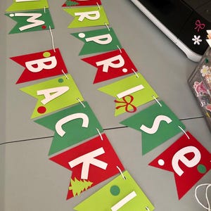 May include: A festive banner with red and green pennants spelling out "SURPRISE BACK!" in white letters. The banner includes decorative elements like a bow and a Christmas tree. The banner is strung with white string.