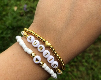 May include: A white beaded bracelet with gold beads and a heart charm. The bracelet spells out the word "AUNTIE".