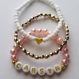 May include: Three bracelets with white, gold, and pink beads. The bottom bracelet has white beads with the name "CARSYN" spelled out in gold letters.
